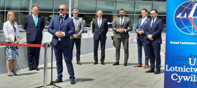 Janusz Janiszewski przemawia na tle terminalu Lotniska Chopina. Za nim stoją pozostali uczestnicy konferencji.